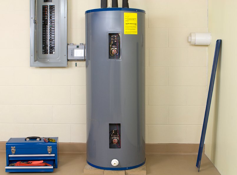 Gray tank water heater in a basement, with electrical panel on the wall to the left. Blue toolbox on the ground to the left.